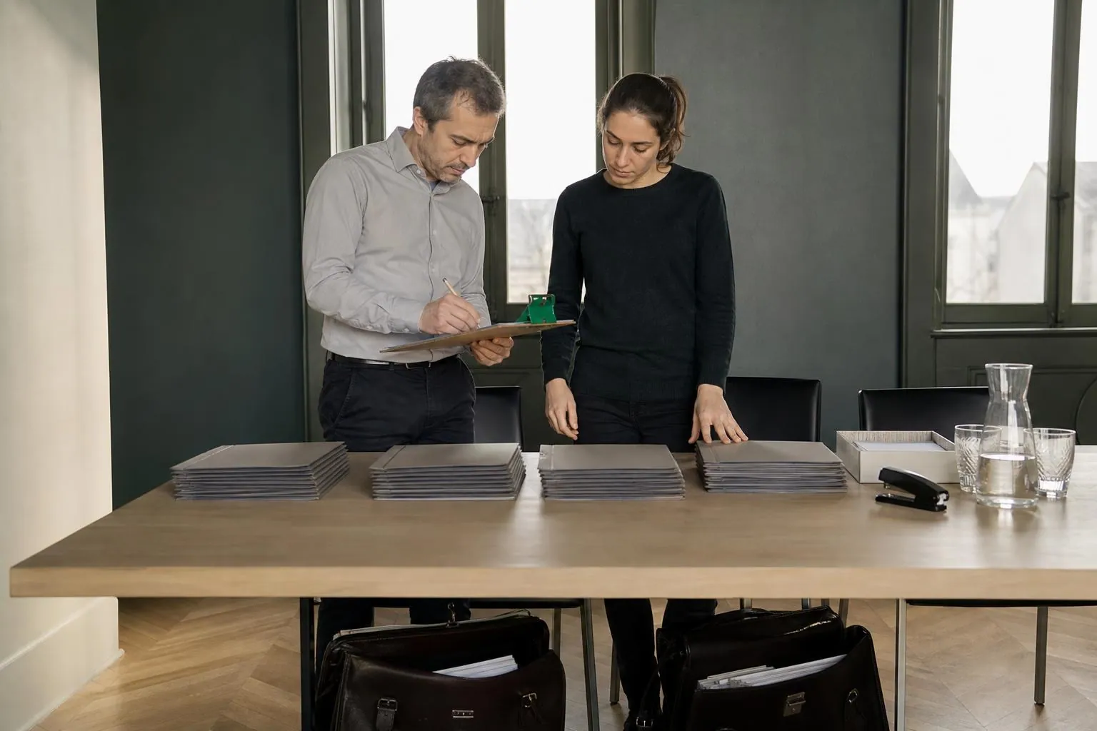 Deux professionnels debout devant une table de réunion cochant à la main une liste posée sur une planchette à pince, sacoches de dossiers au sol