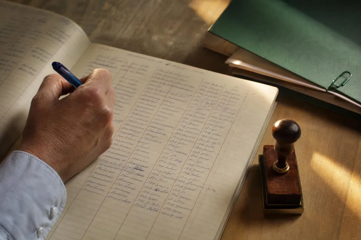 Gros plan sur une page de registre à colonnes remplie à la main, un cachet encreur et une chemise contenant des pièces jointes